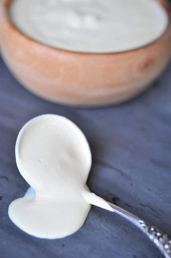 The ultimate vegan cheese sauce dripping off of silver spoon on a slate surface with a wood bowl full of the cheese.