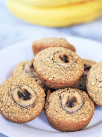A stack of banana muffins on a white plate with bananas in the background