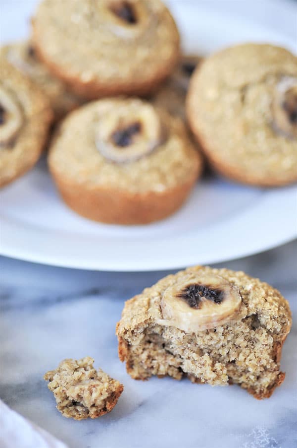 Healthy banana muffins piled on a white plate with one half eaten muffin on the marble surface