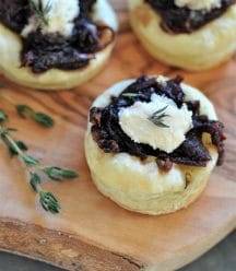 A close up of an onion confit tartlet on a wood board with a sprig of thyme.