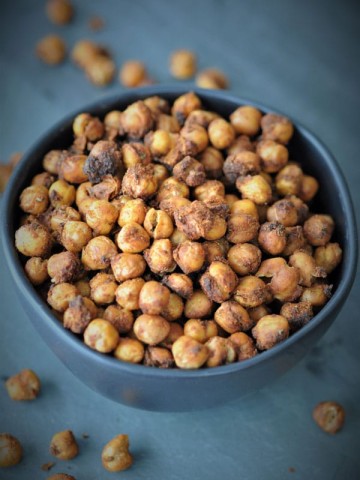 Roasted spicy chickpeas in a black bowl on a piece of slate with spilled chickpeas around the bowl.