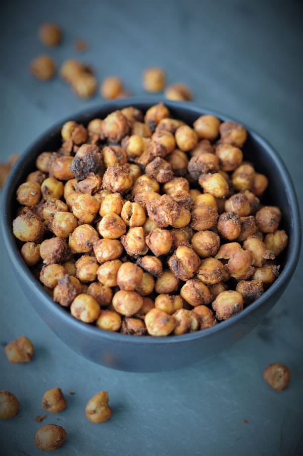 Roasted spicy chickpeas in a black bowl on a piece of slate with spilled chickpeas around the bowl. 