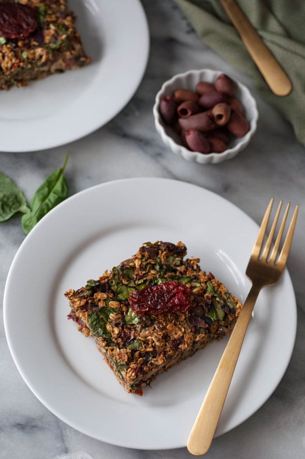 A slice of baked savory oats on a white plate with a gold fork and basil leaves and a dish of kalamata olives in the background