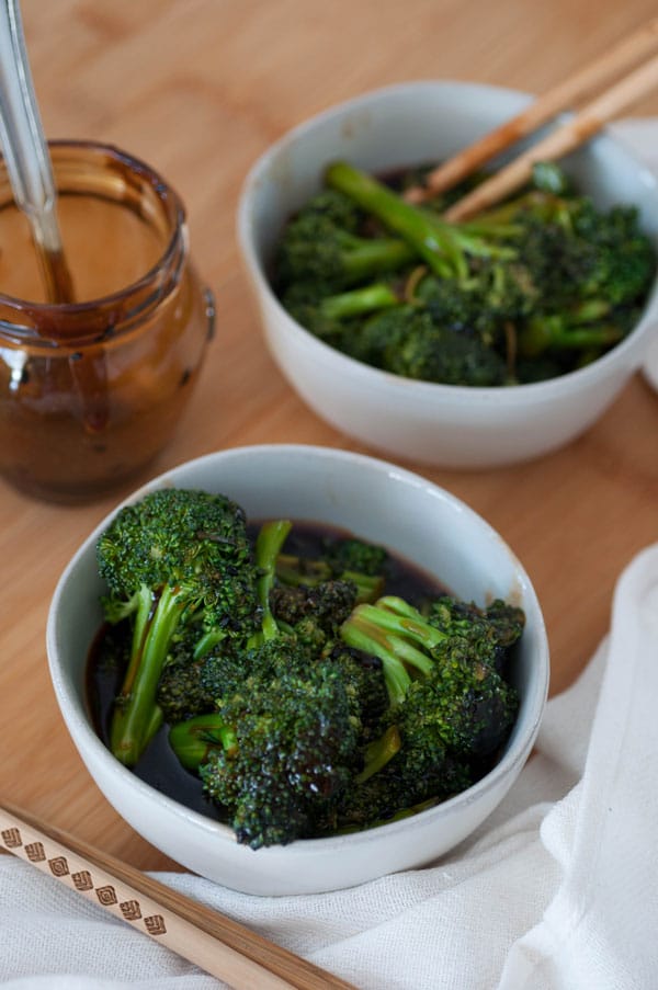 2 white bowls of Teriyaki Broccoli on a wood table with an empty jar of teriyaki sauce with a spoon in it. 