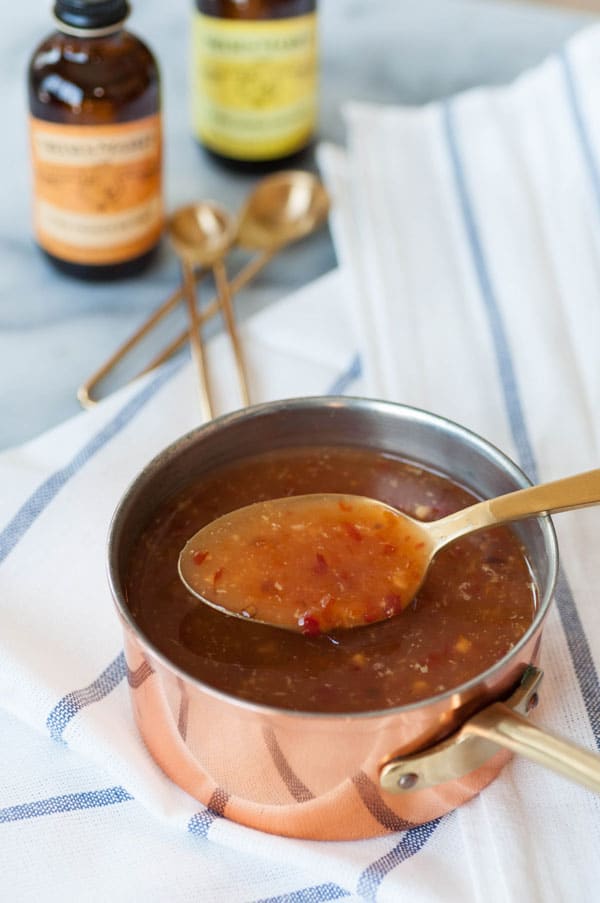 Spicy orange sauce in a copper pan with a gold spoon, sitting on a white and blue striped towel with Nielsen-Massey orange and lemon extract in the background.