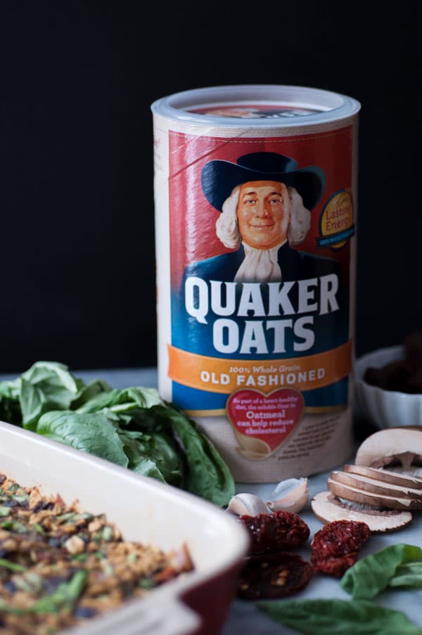 A container of Quaker Old Fashioned Oats with sliced mushrooms, fresh basil, 2 cloves of garlic, and a pan of savory vegan baked oatmeal next to it