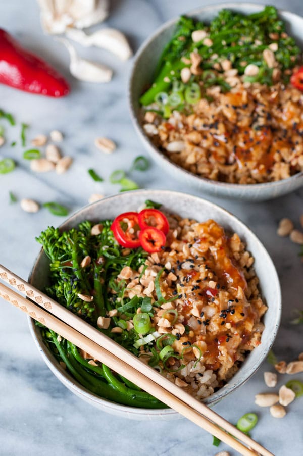 Two bowls of vegan orange chik'n rice bowls with chopsticks and peanuts, chopped green onion, a red pepper, and garlic on a marble counter