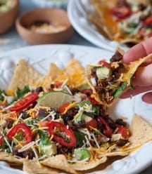 A hand holding a nacho from a white bowl of vegan nachos with another nacho bowl and a wood bowls of vegan cheese, and salsa in the background