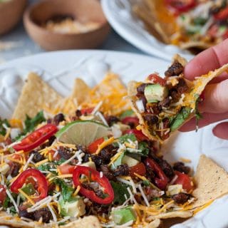 A hand holding a nacho from a white bowl of vegan nachos with another nacho bowl and a wood bowls of vegan cheese, and salsa in the background