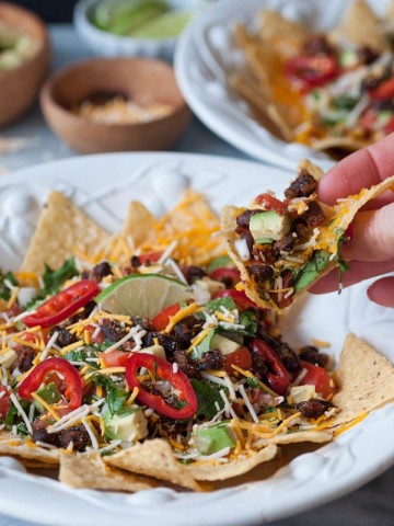 A hand holding a nacho from a white bowl of vegan nachos with another nacho bowl and a wood bowls of vegan cheese, and salsa in the background