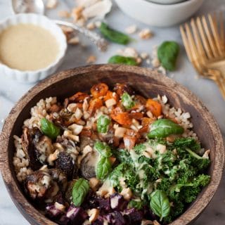 Roasted beets, squash, Brussels sprouts, and kale over brown rice in a wood bowl with chopped cashews and basil. Maple mustard dressing is on the side in a white dish with a stack of white bowls and gold forks on a marble counter.