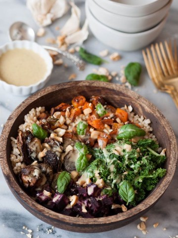 Roasted beets, squash, Brussels sprouts, and kale over brown rice in a wood bowl with chopped cashews and basil. Maple mustard dressing is on the side in a white dish with a stack of white bowls and gold forks on a marble counter.