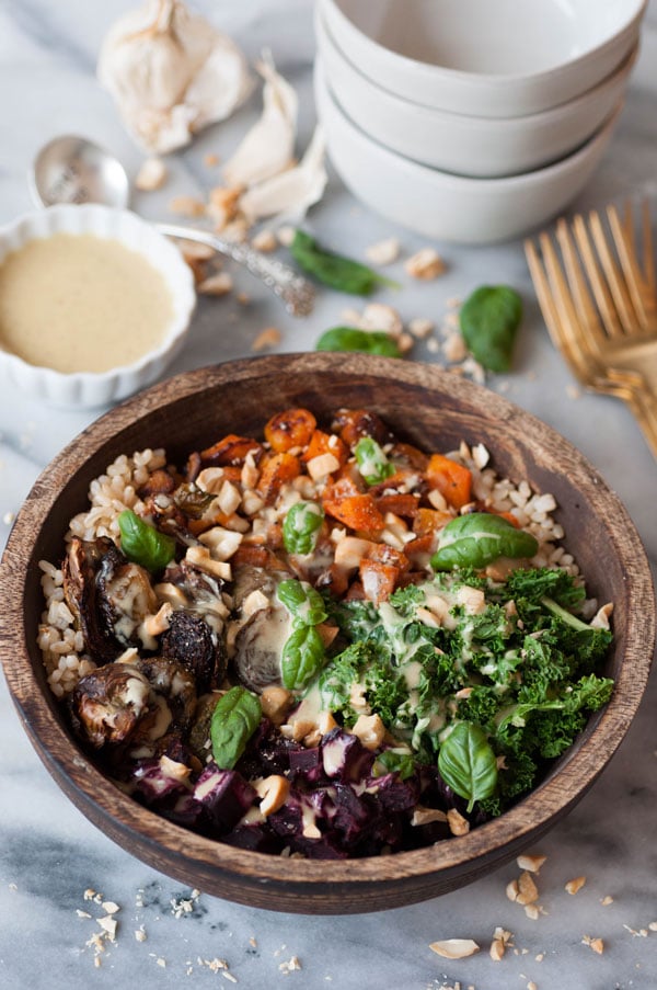 Roasted beets, squash, Brussels sprouts, and kale over brown rice in a wood bowl with chopped cashews and basil. Maple mustard dressing is on the side in a white dish with a stack of white bowls and gold forks on a marble counter.