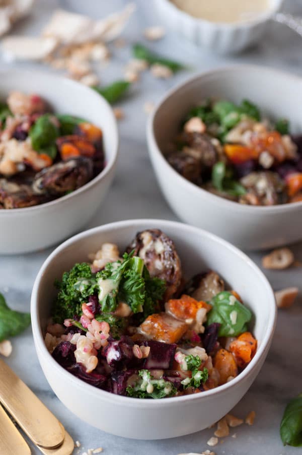 Three white bowls filled with roasted winter vegetables and rice, with three gold forks and a dish of maple mustard dressing in the background.