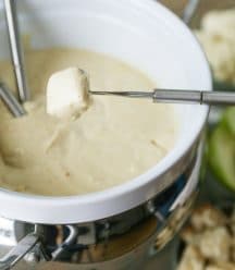 A white ceramic fondue pot filled with golden vegan cheese with a fondue fork dipping a piece of bread into the cheese