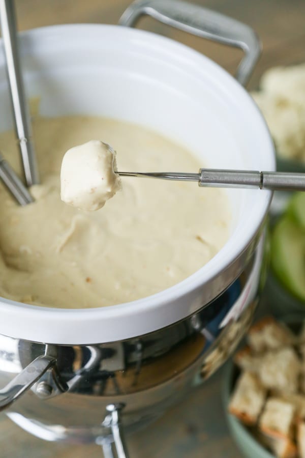 A white ceramic fondue pot filled with golden vegan cheese with a fondue fork dipping a piece of bread into the cheese