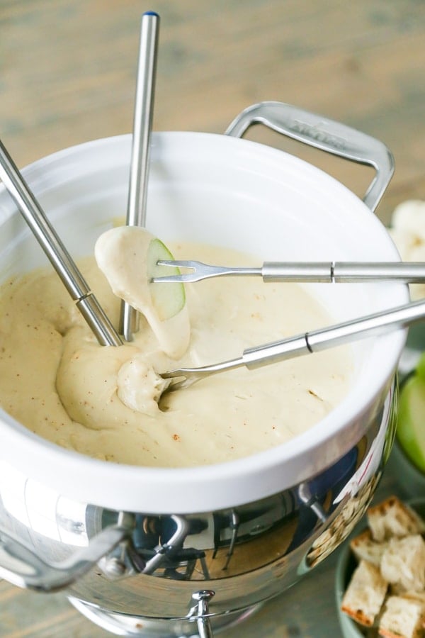 A white fondue pot filled with vegan cheese fondue with four silver fondue forks in the pot, one with a cube of bread and one with an apple slice, the other two are in the cheese