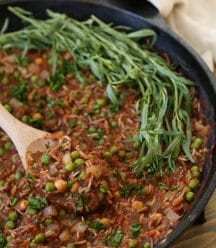Tomato & Rice stew with chickpeas, peas, and Indian spices in an Iron skillet with a wooden spoon and fresh tarragon leaves on one side of the pan