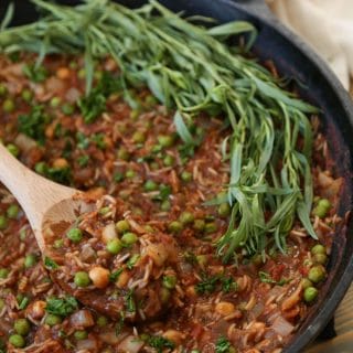 Tomato & Rice stew with chickpeas, peas, and Indian spices in an Iron skillet with a wooden spoon and fresh tarragon leaves on one side of the pan