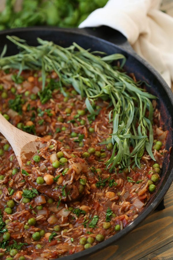 Tomato & Rice stew with chickpeas, peas, and Indian spices in an Iron skillet with a wooden spoon and fresh tarragon leaves on one side of the pan