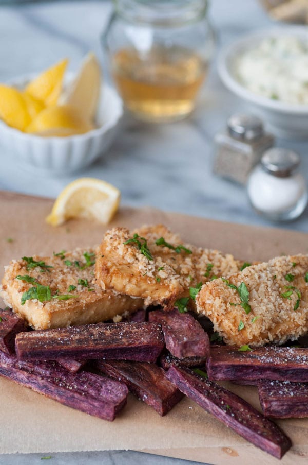 Vegan fish sticks and purple sweet potato fries on a wooden board with a partially squeezed lemon wedge. A bowl of lemon wedges, a jar of vinegar, and a bowl of tartar sauce and salt and pepper shakers are in the background.