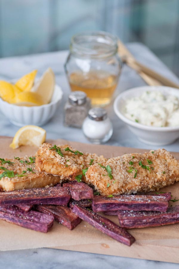 Oven fried vegan fish sticks with purple sweet potato fries on a piece of parchment on a wood board with tarter sauce, lemons, vinegar, and salt and pepper in the background