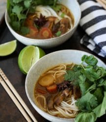 Two bowls of shiitake pho in white speckled bowls garnished with fresh cilantro and basil with chopsticks and lime wedges and a blue and white striped towel next to them