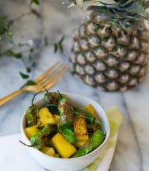 Shishito Peppers with pineapple in a white bowl with a gold fork and half a pineapple in the background