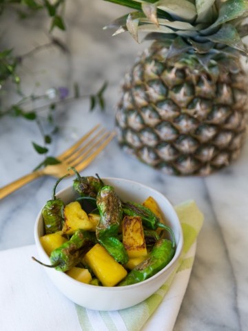 Shishito Peppers with pineapple in a white bowl with a gold fork and half a pineapple in the background