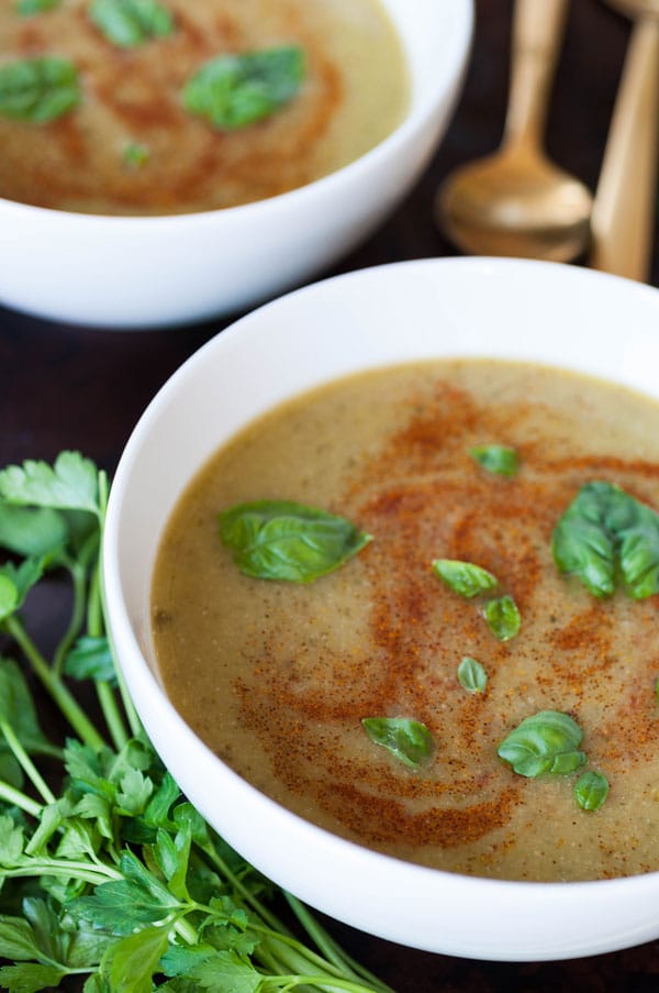 Two white bowls full of spicy green soup with basil leaves and paprika swirls and 2 gold spoons and parsley leaves on the sides of the bowls