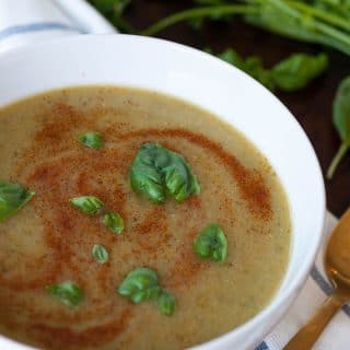 Spicy green soup in a white bowl with basil leaves and swirls of paprika on top. Sitting on a blue and white striped towel with a gold spoon and parsley and lemon on the side.
