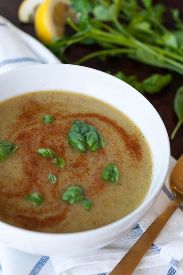 Spicy green soup in a white bowl with basil leaves and swirls of paprika on top. Sitting on a blue and white striped towel with a gold spoon and parsley and lemon on the side.