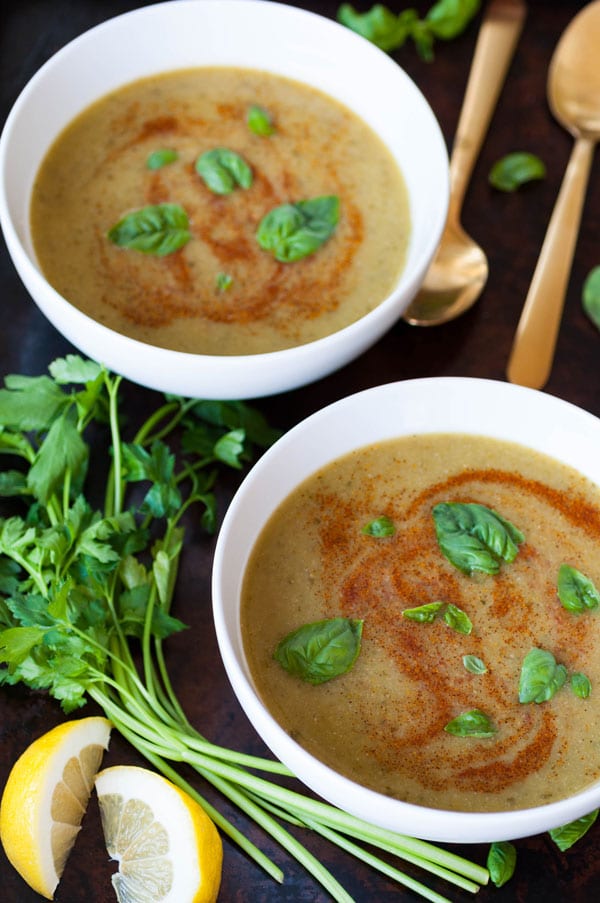 Two white bowls of spicy green soup with basil and paprika swirls on a dark surface with 2 gold spoons and parsley sprigs and lemon wedges on the side