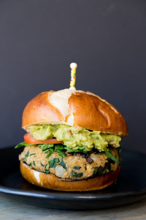 A thick spinach burger patty between a pretzel bun, shredded fresh spinach, sliced tomato, and mashed avocado on a black plate with a yellow and red toothpick stuck on the top of the bun