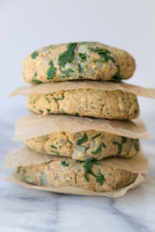 Four uncooked spinach burgers stacked between parchment paper on a marble surface