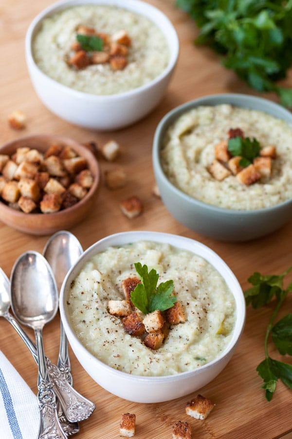 3 bowls of Colcannon potato & cabbage soup, 2 in white bowls and the middle bowl is green, with 3 silver spoons and a wood bowl with croutons and more croutons and parsley sprinkled on the wood table.