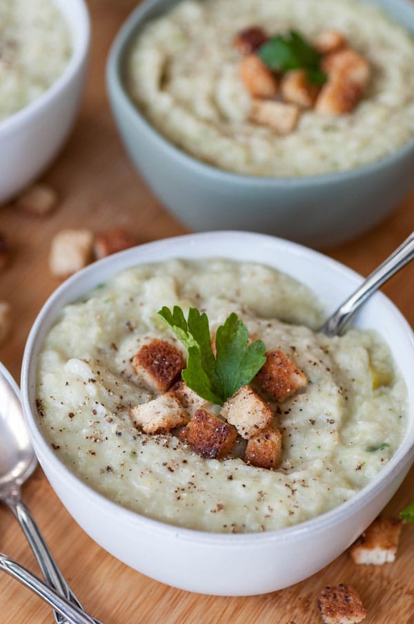 Potato & cabbage soup in a white bowl with croutons and a sprig of parsley on top with a silver spoon in the soup. Two silver spoons and two more bowls of soup are in the background.