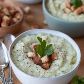 A bowl of potato & cabbage Colcannon soup with croutons and a sprig of parsley and two more bowls and a wood bowl of croutons and silver spoons in the background.