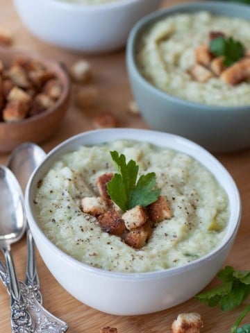 A bowl of potato & cabbage Colcannon soup with croutons and a sprig of parsley and two more bowls and a wood bowl of croutons and silver spoons in the background.