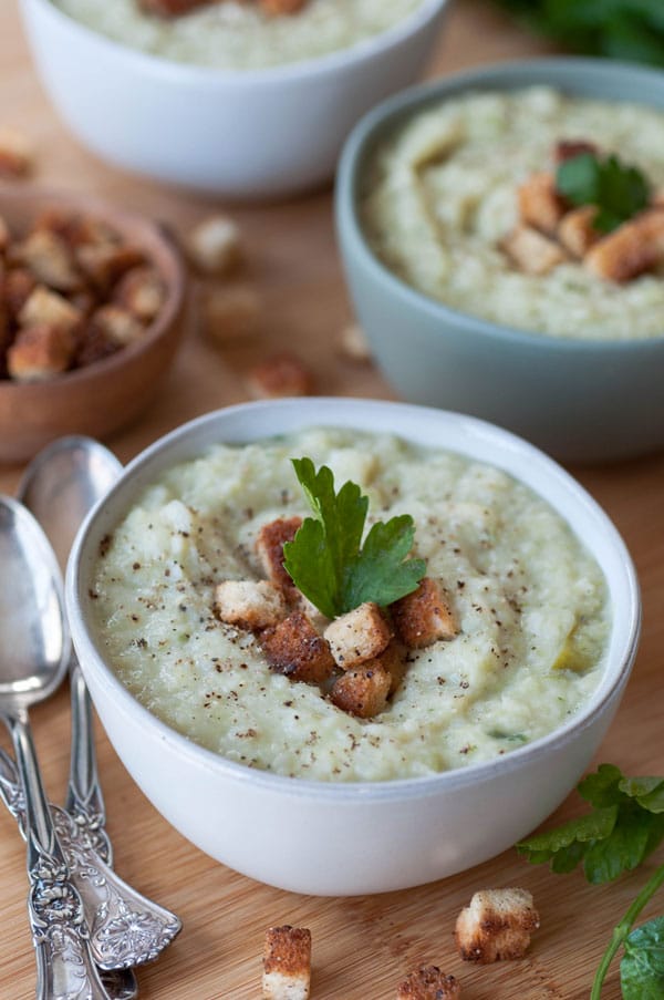 A bowl of potato & cabbage Colcannon soup with croutons and a sprig of parsley and two more bowls and a wood bowl of croutons and silver spoons in the background. 