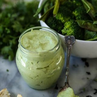 A jar of green goddess dressing with a small silver spoon of dressing leaning on a white bowl of salad, and two pieces of bread in front of it with a pile of parsley in the background