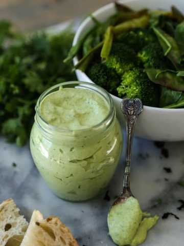 A jar of green goddess dressing with a small silver spoon of dressing leaning on a white bowl of salad, and two pieces of bread in front of it with a pile of parsley in the background