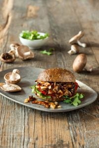 A shiitake BBQ sandwich featured in the Veganosity cookbook Great Vegan BBQ Without a Grill on a gray plate with whole shiitake mushrooms and a bowl of greens in the background