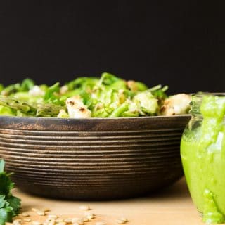 Green Spring Vegetable salad in a wood bowl on a wood surface with sunflower seeds sprinkled in front and part of a glass jar of creamy chimichurri sauce