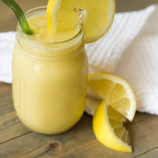 A mason jar filled with vegan lemon slushy with a slice of lemon on the glass and a green straw with lemon wedges and a white napkin on a wood table