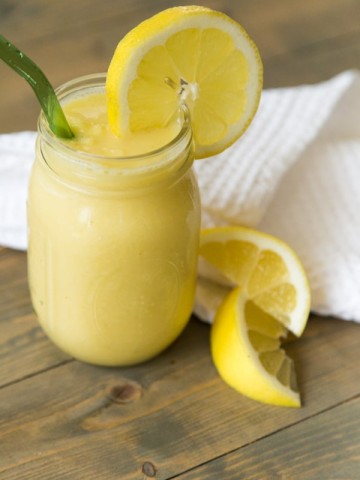 A mason jar filled with vegan lemon slushy with a slice of lemon on the glass and a green straw with lemon wedges and a white napkin on a wood table