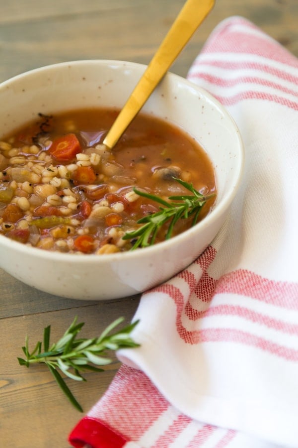 Italian Lemon Rosemary Barley Soup next to a white and red napkin with rosemary and on a wood cutting board