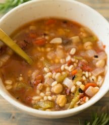 A white bowl full of Italian Lemon Rosemary Barley Soup with a gold spoon in the bowl and sprigs of rosemary around it on a wood table