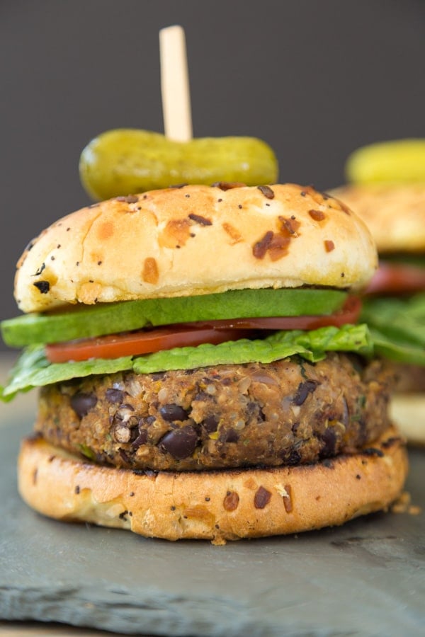 A black bean burger with lettuce, tomato and avocado slices on an onion bun with a pickle on top with a toothpick