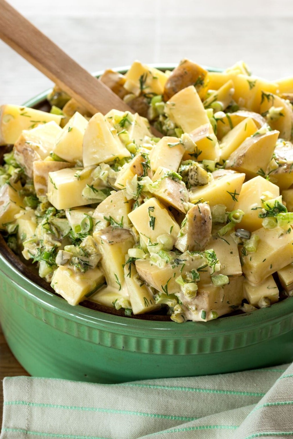 A close up of vegan potato salad in a green bowl with a wooden spoon sticking out of it